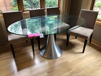 Overall view of oval glass dining table with four upholstered chairs arranged around it in a room with wooden floor and large windows.