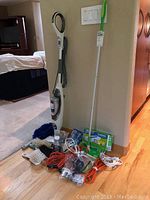 Full view of Shark steam cleaner, Swiffer mop with refills, assorted brushes, extension cords, and cleaning accessories arranged on the floor near a wall.