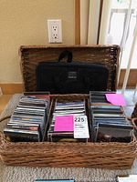 Front view of large woven basket containing over 50 jewel case CDs and a black fabric lined carrying case with handle inside basket.