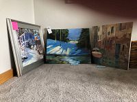 Three acrylic paintings displayed on floor. Left one framed with silver frame depicting snowy scene. Middle one shows snowy forest landscape. Right one depicts canalside scene with boats.