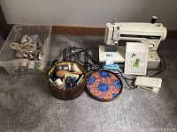 Full lot overview showing the Kenmore sewing machine, accessories in plastic container, wooden basket with sewing supplies, and a round fabric patchwork item with pincushion.