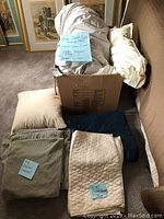 Full bedding lot including a cardboard box with duvets and pillows, beige king coverlet folded, and cream silk pillow sham laid out.