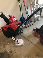 Front three-quarter view of Toro PowerClear 210E snow blower showing red housing, black handle and white wheels with operator manual on floor