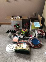Shows the full assortment of holiday items laid out on carpet including two cardboard boxes with ornaments, paper plates, napkins, ceramic plate, bead garland, and various other decorations.