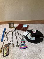 Photo showing six necklaces with various pendants including a carved figure, glass heart, colorful fabric, and jade-style pendants, along with 8x8 glass plate, trinket box, and display stand on carpeted floor.