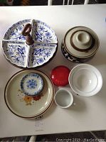 Image showing assorted serving ware including blue and white divided serving stand, brown pottery jar with lid, floral plate, small pitcher, white bowl, red dish and small blue floral bowl.