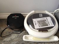 Photo of two George Foreman grills; one black rectangular classic model and one white circular model with clear dome lid and manual on top