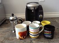 Bosch Tassimo coffee maker, glass French press, and six assorted coffee mugs placed on a floor surface against a wall.