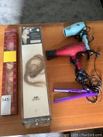 Photo showing two hair dryers, purple flat iron, and two packages of hair extensions on a wooden surface.