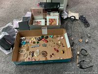 Photo of assorted costume jewelry laid out in cardboard box and on floor, including colorful brooches, earrings, chains, and watches.