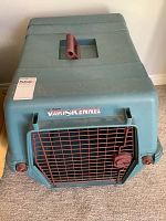 Blue plastic Doskocil Deluxe Vari Kennel dog crate with red metal door, includes a handle on top and ventilation slots.