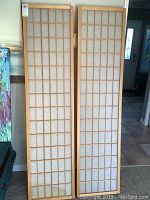 Two 3-panel wood framed folding screens with fabric inserts leaning against a wall, showing the grid pattern and overall condition.