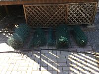 Five rolls of green plastic coated chain link fencing, each approximately 36 inches high, rolled and laid side by side on a paved surface. Also includes a 12 foot watering hose with three sprinkler heads attached shown across the front.