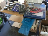 Overview of the lot items on and around a small table including four round lights, socket wrench set, cable roller, and shelf unit.