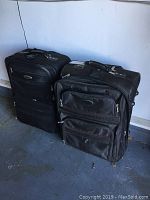 Two black soft-sided suitcases side by side on garage floor, showing front and top views highlighting zippered pockets and handles
