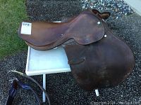 Photo of the full brown leather English riding saddle on a white table outdoors with visible grass and stones nearby. Saddle shows moderate signs of wear.