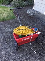 True Temper CD20 red lawn spreader with black handle, wheels, loaded with yellow and blue extension cords and white power strip inside.