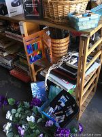 Full view of wooden bookshelf with multiple shelves containing art supplies like paints, coloring set, baskets and prints, plus some stacked items