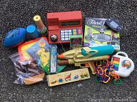 Photo showing all items in the lot including the red Kids Learning Resources cash register, Super Soaker gun, wooden puzzle, colorful plastic toys, and stacked cups.