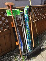 Photo showing the full assortment of the garden tools leaning against a wooden fence outdoors, including the beach umbrella, snow shovel, Swiffer mop, garden rake, regular umbrella, and Fiskars weed puller.