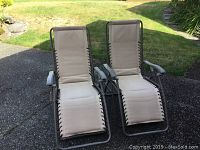 Photo of two beige folding lounge chairs with gray metal frames, showing the front view and overall condition including fabric and armrests.
