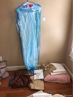 Full view of bedding items, showing packaged item, blankets, and folded bed linens on floor and leaning against wall.