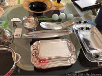 Photo showing an asymmetrical silverplate bowl, elongated silver trays, a rectangular silverplate tray with decorative handles, etched pewter serving dish, and small decorative round items on a glass table