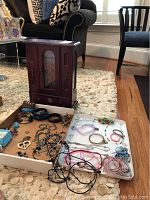 Wooden jewelry box with glass door and drawer, several costume jewelry pieces displayed in boxes and trays placed on beige carpet.