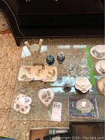 Overview of lot items on glass table including porcelain dishes, vases, atomizers, clock, flower air freshener.