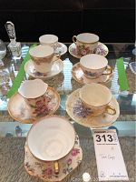 Eight porcelain tea cup and saucer sets with floral patterns and gold accents arranged on glass table
