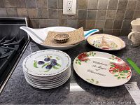 Overview showing all plates and serving items arranged on kitchen counter: appetizer plates with plum design stacked, Christmas plate with floral ribbon design, Eiffel Tower plate, large white bowl with built-in salad forks, small serving plates with dip bowls.