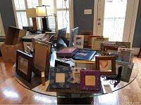Wide view of many assorted picture frames including wicker style, floral, leopard print, wooden, and pewter, along with two photo albums on a glass table.