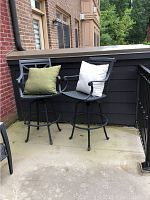 Pair of black metal outdoor bar stools placed on a concrete patio. Each stool has a cushion, one green and one white.