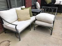 Outdoor metal frame loveseat and chair with white cushions and two accent pillows against brick wall and patio