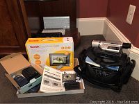 Group view of all electronics on carpet: two Canon camera boxes, Kodak picture frame box, Sony camcorder on bag, keyboards on shelf