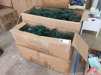 Cardboard boxes labeled Frontgate Holiday Collection with artificial greenery visible inside representing the holiday trees and garland set.