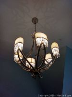 Photo of the chandelier lit, showcasing six cream shades with hanging crystal beads attached to each shade, mounted on curved metal arms.