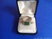 Gold ring with rectangular green jade center displayed in a black velvet presentation box against blue background
