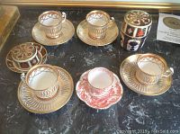 Five cups and saucers on black marble surface, four white with gold ornate patterns and one red and white with bird design
