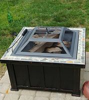 Photo of the black metal Hampton Bay fire pit with tile frame top, removable mesh cover, and firewood inside on a paved surface outdoors.