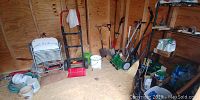 Photo showing folded white metal chair, Milwaukee hand truck, manual reel mower, shovels, and garden buckets inside wooden shed.