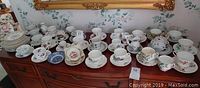 Wide view showing 24 cups, saucers and small plates in various floral patterns and styles on a wooden sideboard.