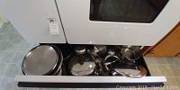View of pots and pans drawer under oven, showing multiple stainless steel pots, pans and lids stacked together.