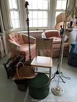 Overview of the room showing three vintage floor lamps, pink fabric lampshade, light wooden chair, leather foot stool, and baskets on carpeted floor near windows.
