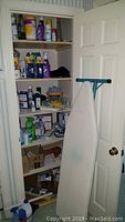 Shot of closet with multiple shelves stocked with cleaning supplies and household items. White ironing board leaning against door.