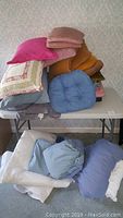 Pillows and cushions stacked on a table, showing various colors and styles including pink, tan, blue, and patterned pillows.