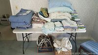 Wide view of table with piles of bed sheets, blankets, pillows, towels, and boxes of hangers underneath.