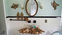 Photo of several gold-toned decorative items mounted on a bathroom wall including an oval mirror, matching ornate candle sconces, cherub motifs, and bird wall plaques under lighting.