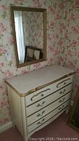 Front angle view of the cream dresser with gold trim and matching wall mirror hung above it, showing the dresser's four drawers with decorative handles.
