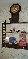 Front view of wooden lattice wall shelf with built-in octagonal clock and three small drawers, displaying various decorative items including Hummel style figurine, yellow toy car, dolphin figurine, bird figurine, horse figurine, and boxed set of Oriental china.